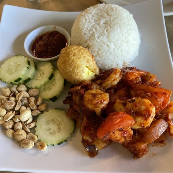 A white plate of Nasi Lemak with chili prawns, rice, fried egg, peanuts, cucumber, and sambal.