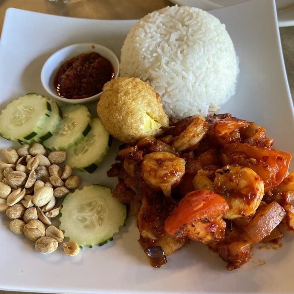 A white plate of Nasi Lemak with chili prawns, rice, fried egg, peanuts, cucumber, and sambal.