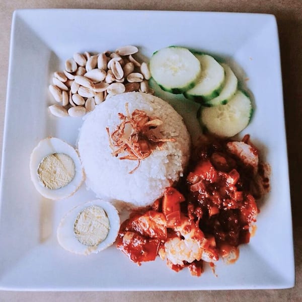 Nasi Lemak with rice, sambal prawns, peanuts, cucumber, and hard-boiled eggs on a white plate.