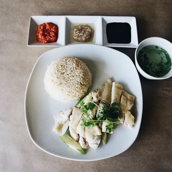 Hainanese chicken rice with sliced chicken, rice, soup, and three dipping sauces on a white plate.