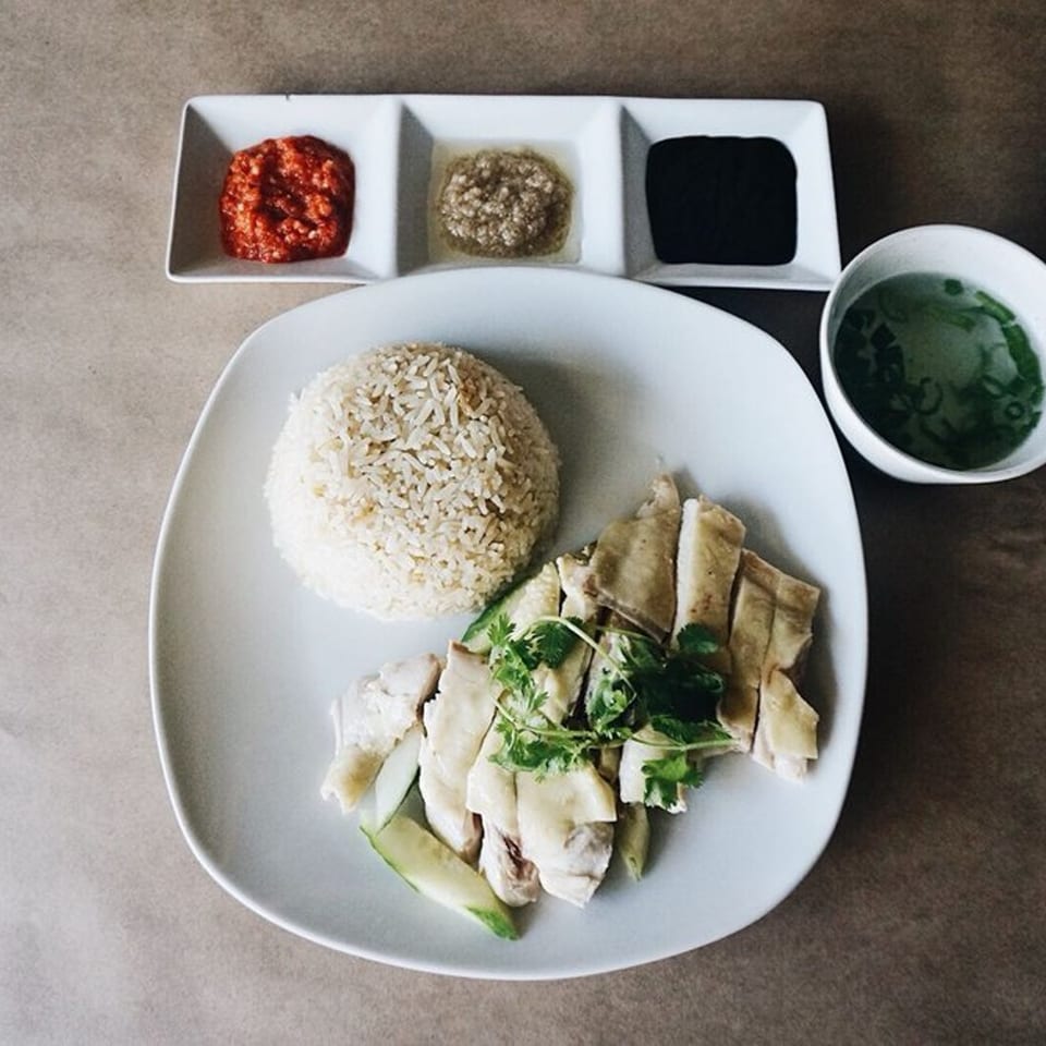 Hainanese chicken rice with sliced chicken, rice, soup, and three dipping sauces on a white plate.
